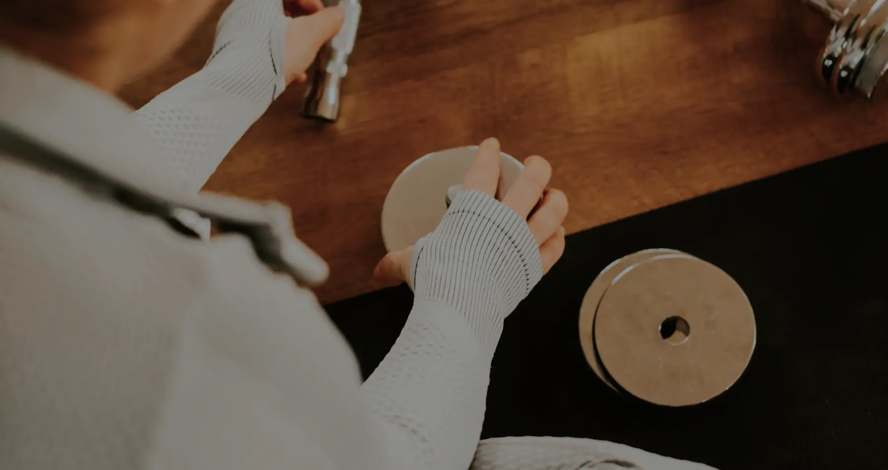 A person wearing white athletic gloves is assembling a pair of dumbbells on a wooden surface, emphasizing a focus on wellness and premium fitness equipment.