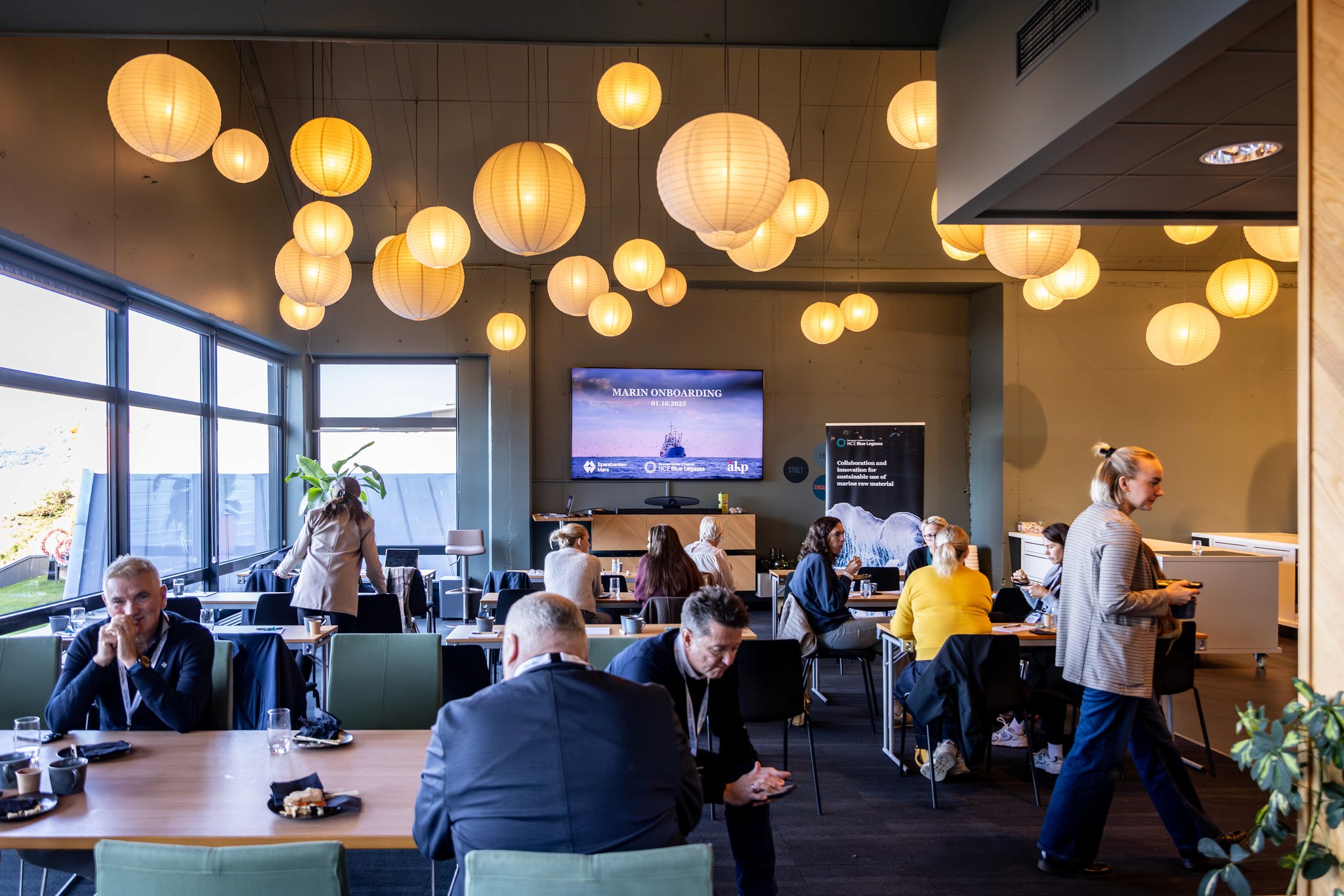 A vibrant restaurant interior with warm lighting and people enjoying meals at tables. A screen is visible in the background.