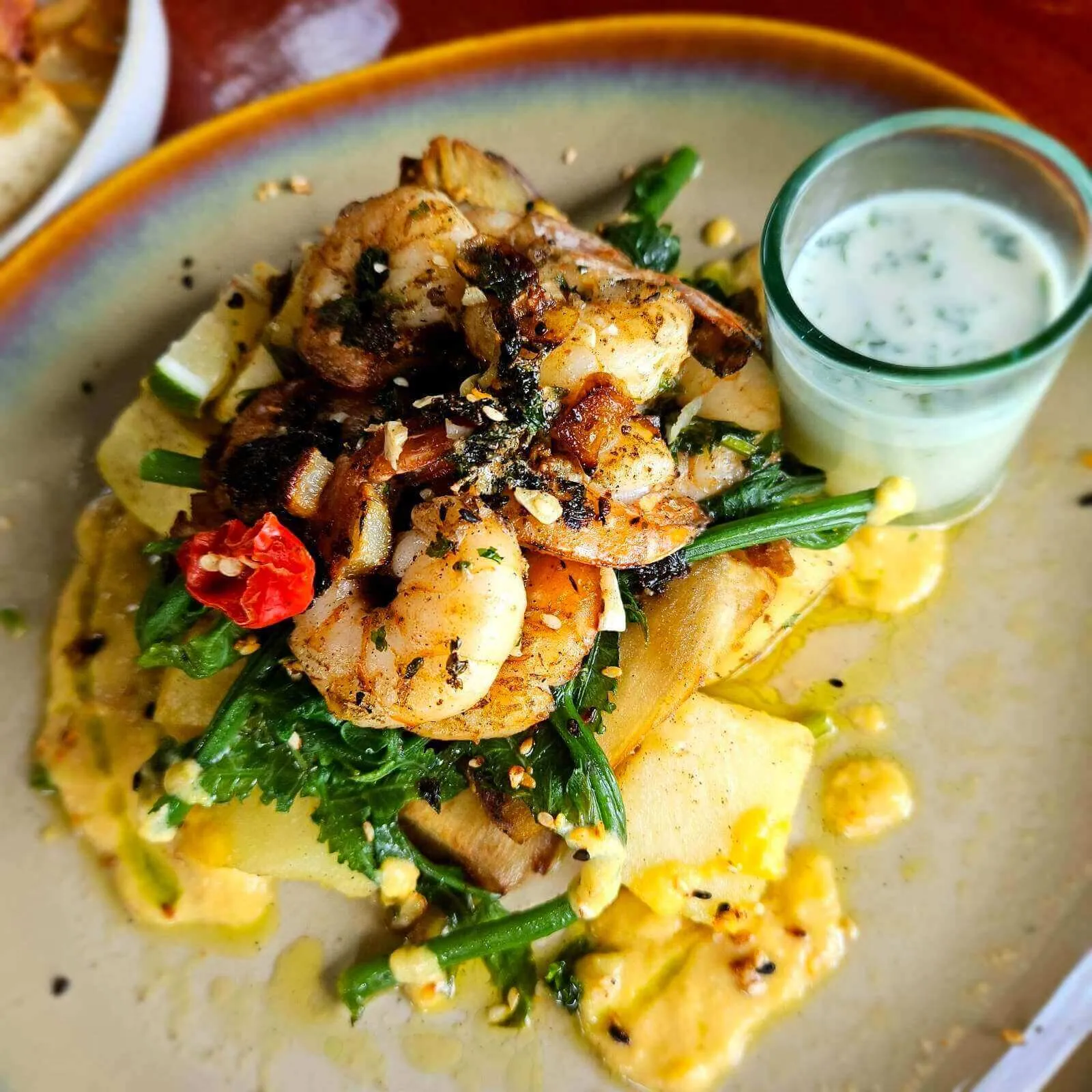 Grilled prawns, sautéed greens, and root vegetables on a plate, with a creamy yellow sauce and Kokoda