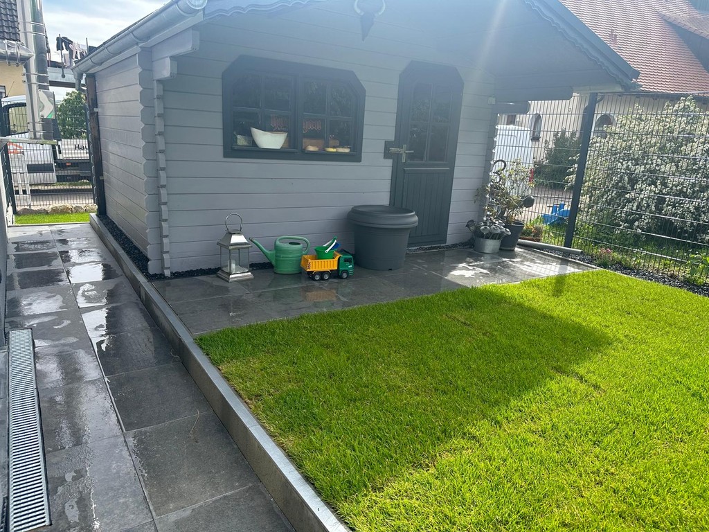 Gartenhaus mit nasser Steinterrasse und frisch verlegtem Rollrasen; Gießkanne, Spielzeug-Laster und Pflanzkübel vor dem Eingang.