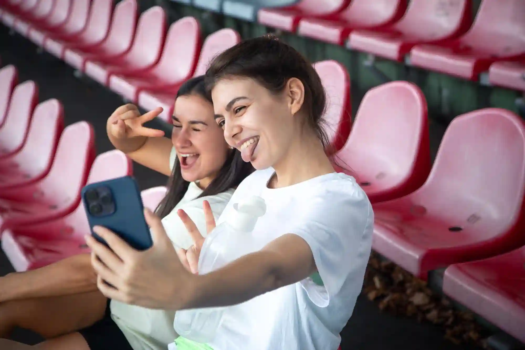 Two young women making fun faces and peace signs while taking a selfie in empty stadium seating.