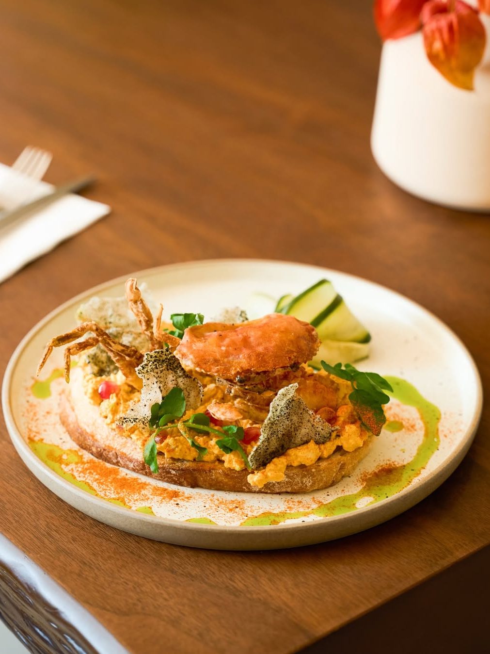 A beautifully presented dish featuring crispy crab, creamy sauce, and garnishes on a plate, accompanied by cucumber slices. The dish is served on a wooden table.