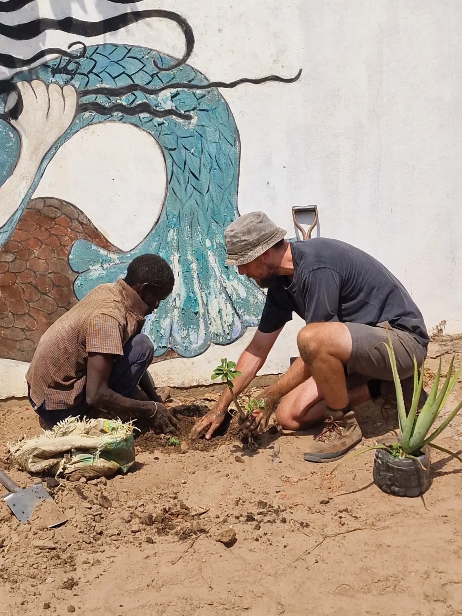 A child and an adult are planting in soil near a colorful mural, engaged in gardening activities together.