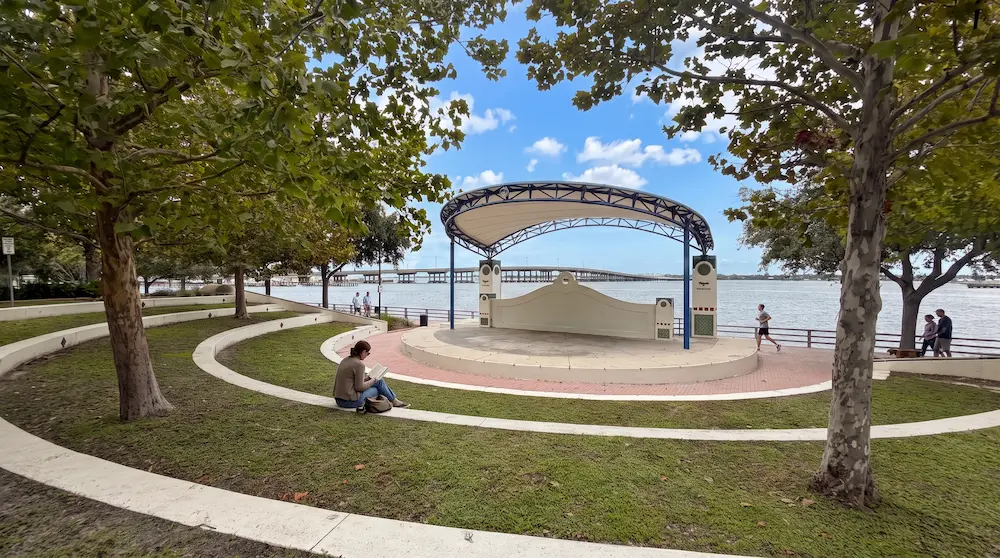 The image features a serene view of the Riverwalk in Bradenton, FL, showcasing a small amphitheater surrounded by lush green trees and curved pathways along the waterfront.