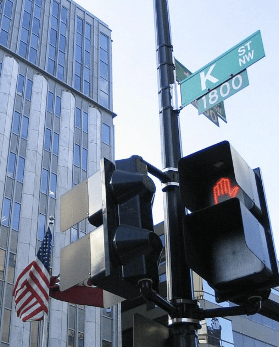 A traffic signal with a red light and a street sign on a building corner against a clear blue sky.