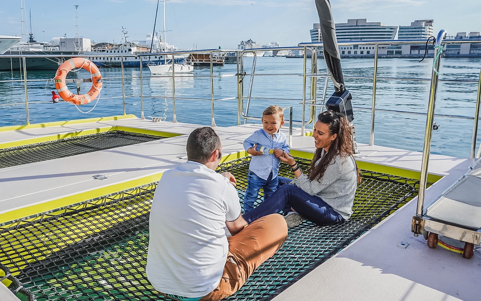Turisti rilassati sul ponte di una crociera in catamarano con un bambino, vista sul porto sullo sfondo.
