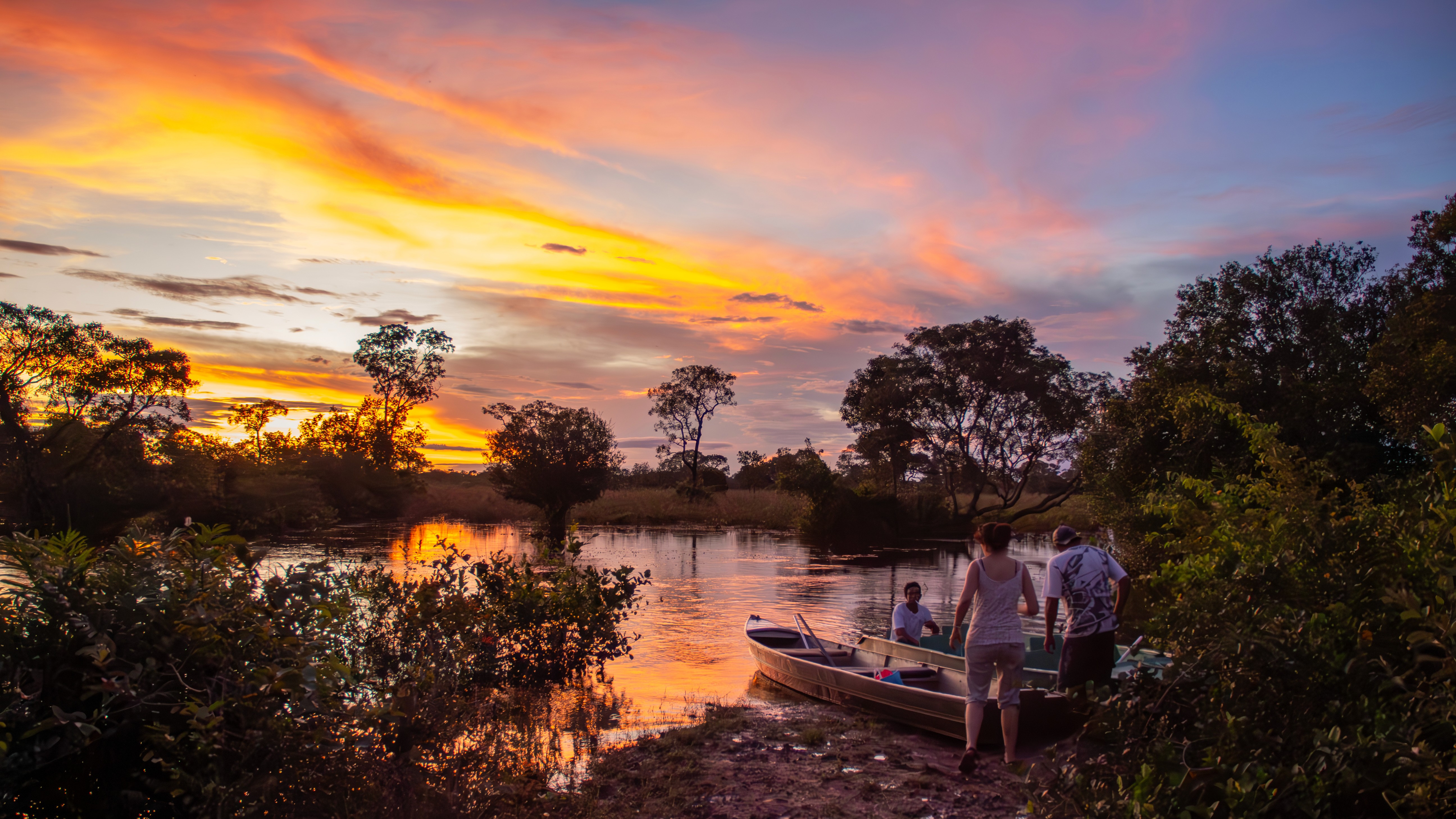 Three people board a small boat at sunset on a quiet river in the Pantanal, surrounded by lush vegetation and the golden reflections of the sky on the water.
