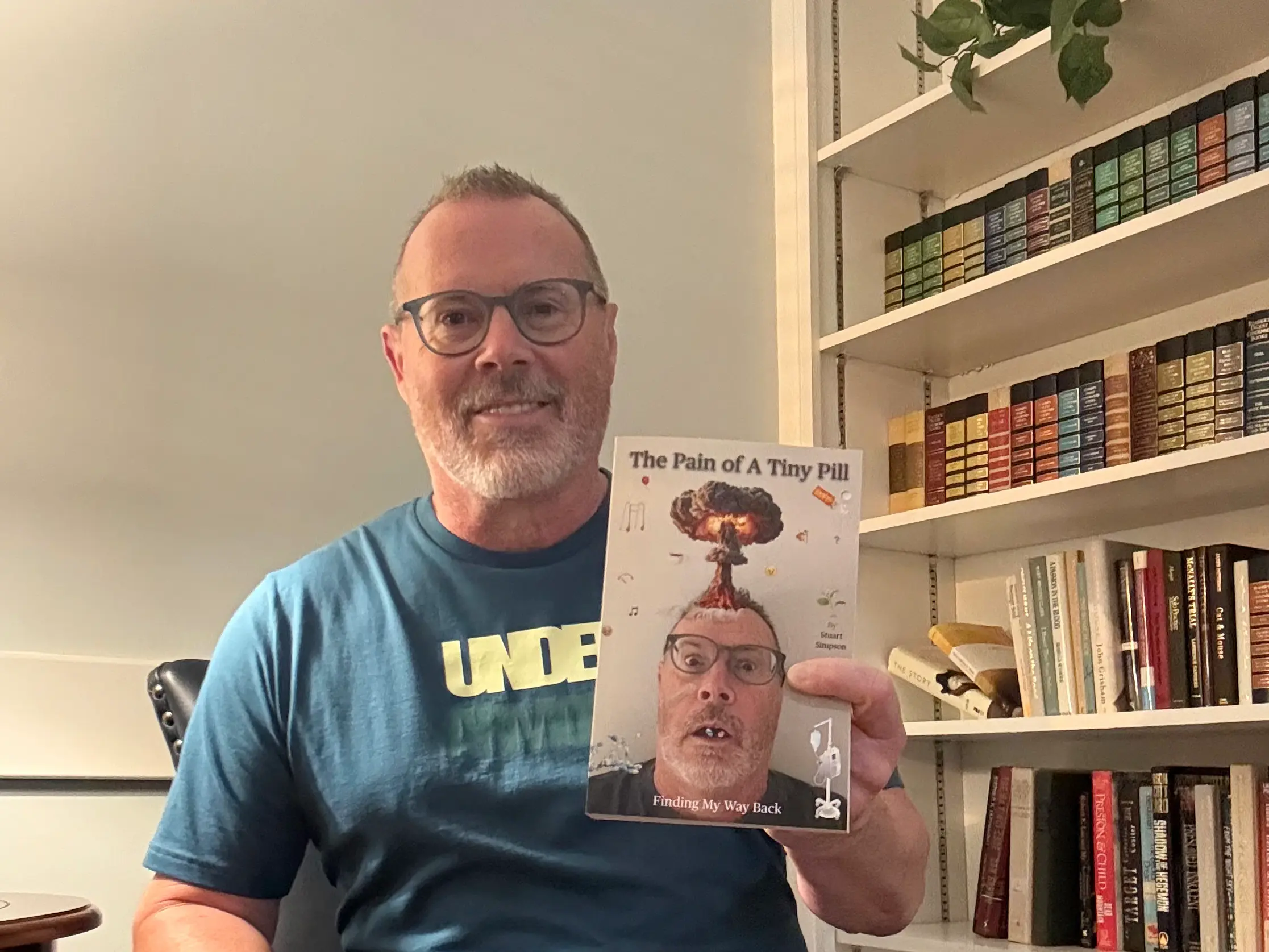 Stu holding a copy of his book “The Pain of a Tiny Pill: Finding My Way Back” in front of a bookshelf