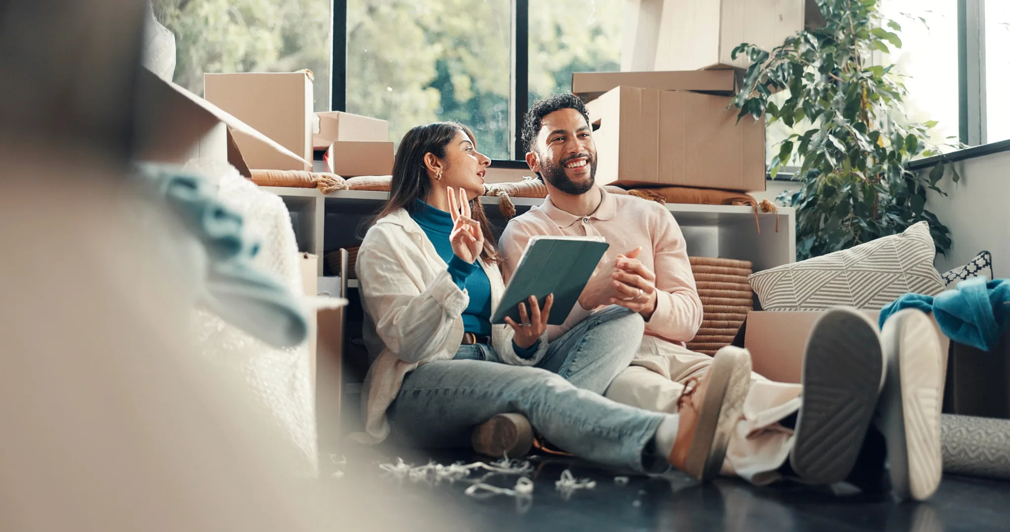 A couple sits on a couch surrounded by cardboard moving boxes, holding a tablet together and smiling while planning their relocation in a bright, plant-filled room.
