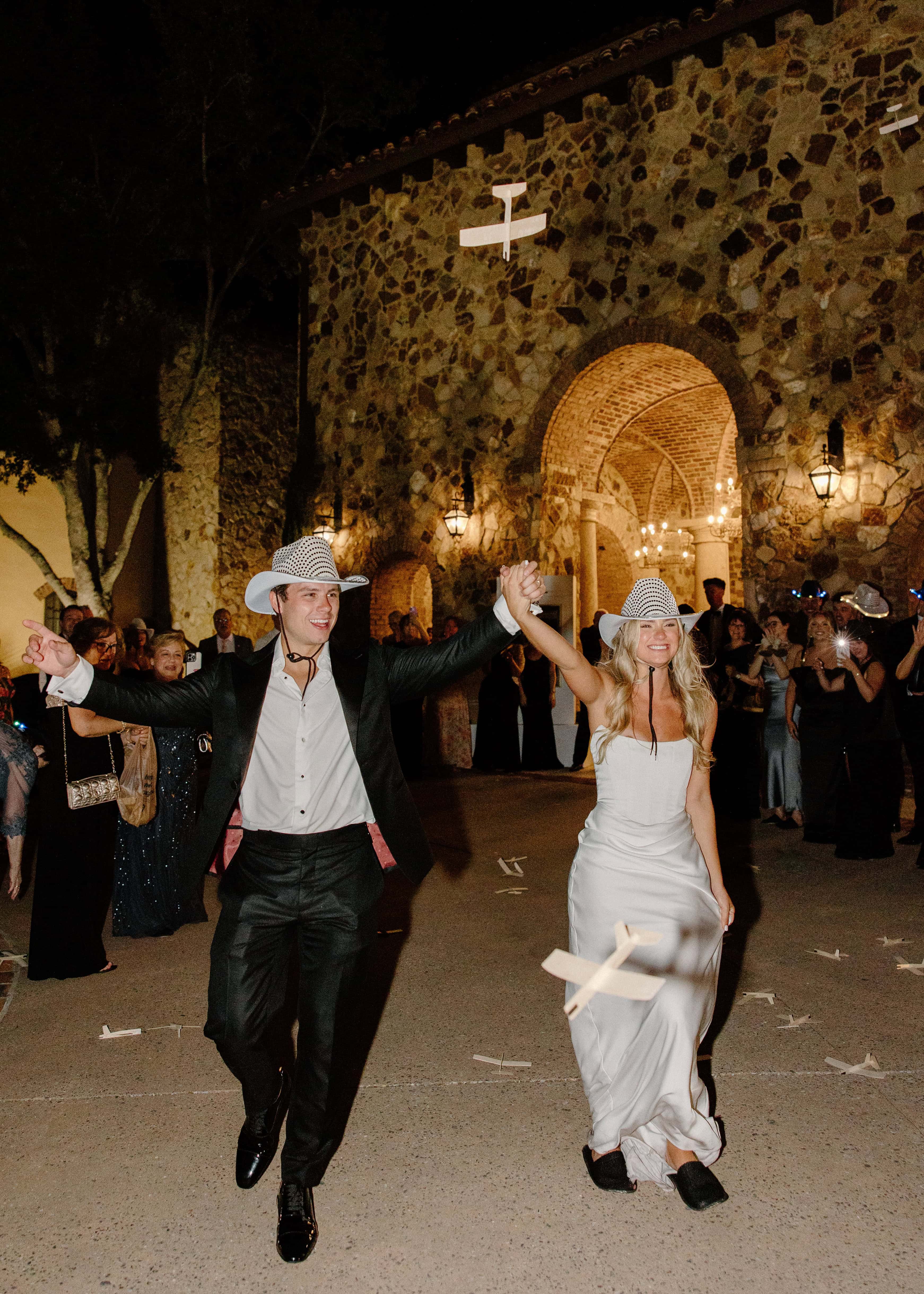 A photo captures a couple walking toward the camera at night, both wearing hats and holding hands. There are people in the background and what looks like confetti on the ground.