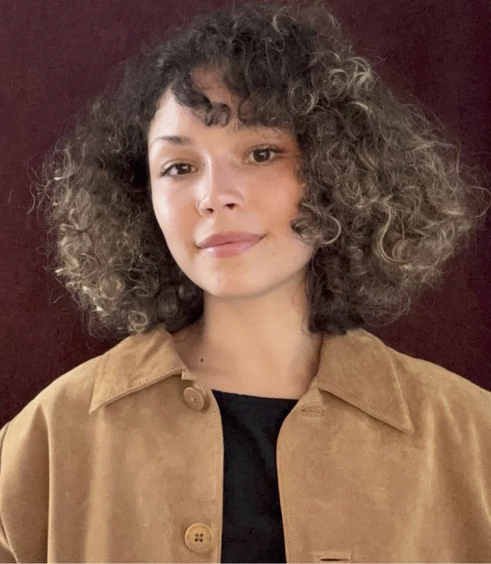 Uma pessoa com cabelo cacheado na altura dos ombros está vestindo uma jaqueta marrom sobre uma camisa preta, em pé diante de um fundo vermelho escuro.