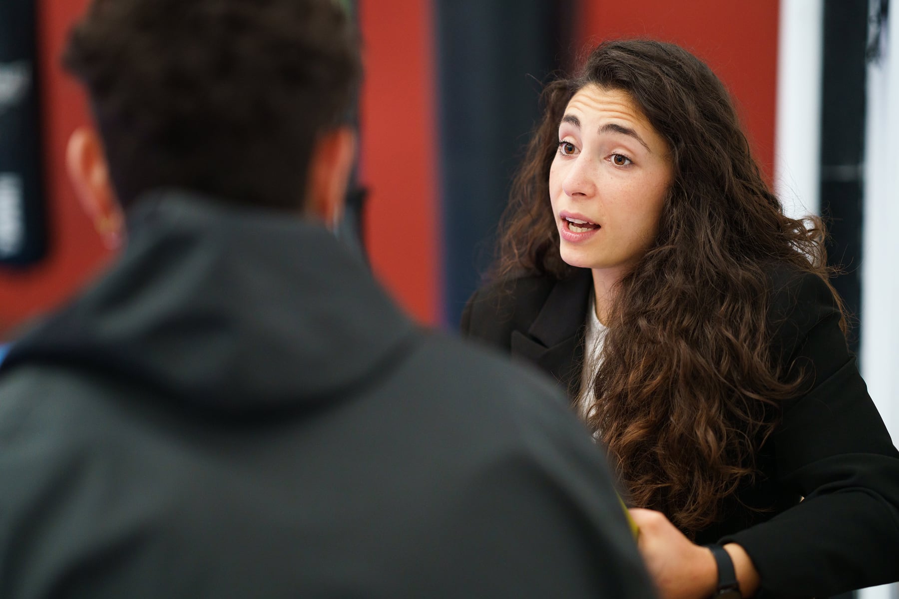 A woman with long curly hair talks to someone across the table during a conversation or interview. The listener is out of focus in the foreground.