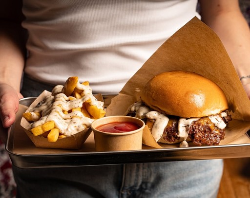 A person holding a tray with a burger, fries, and a small dessert, set against a warm background.