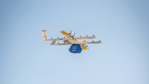 A white and yellow drone in flight against a clear blue sky; it is carrying a walmart package, demonstrating modern delivery technology.