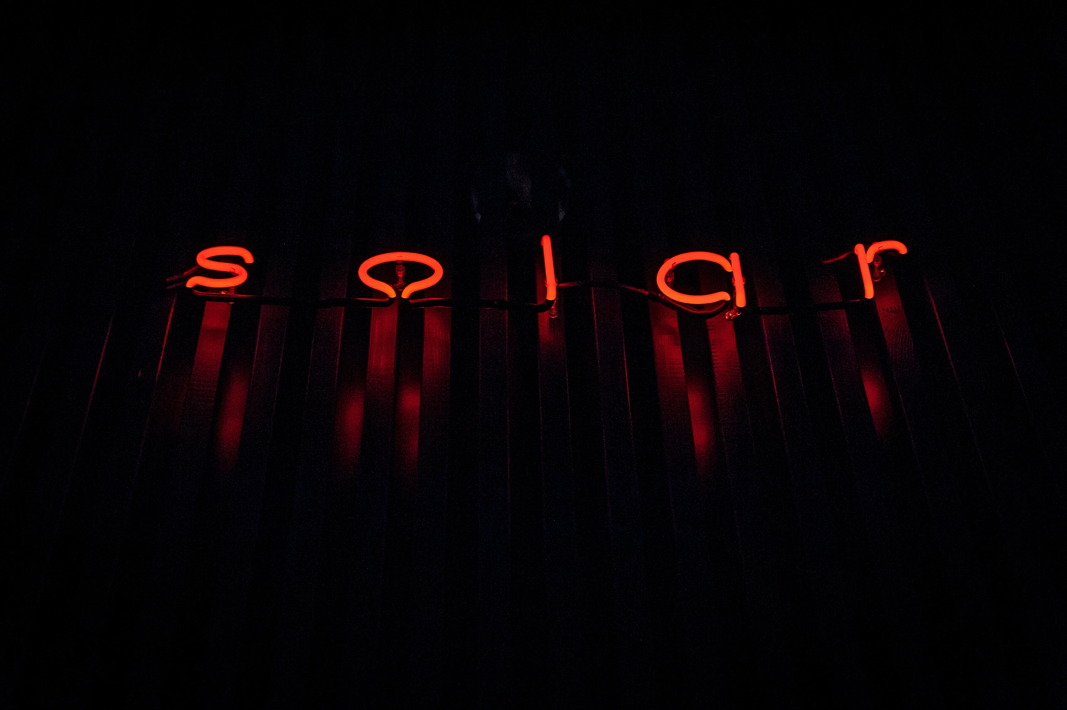 Glowing red neon lettering solar mounted on a dark slatted wall in near-total darkness at SOLAR SKY LOUNGE.