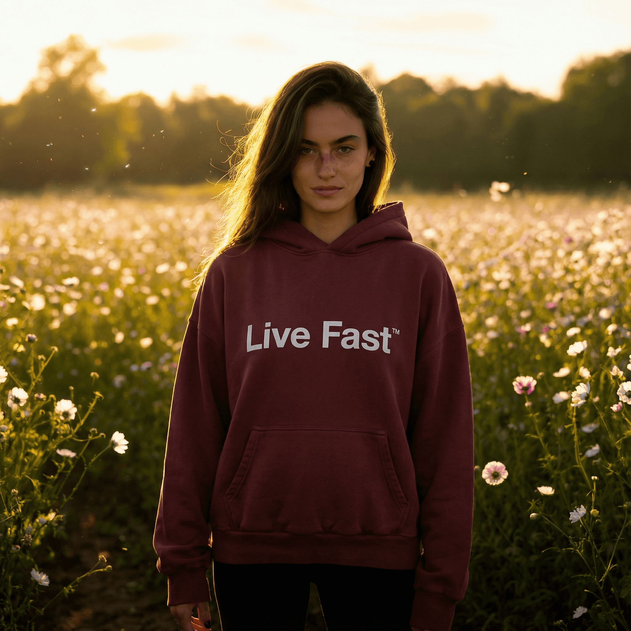 Weibliches KI-Model in burgunderrotem Hoodie mit ‚Live Fast‘-Print, stehend in einem blühenden Feld bei goldenem Sonnenuntergang – ideal für Fashion-, Outdoor- und Lifestyle-Content.