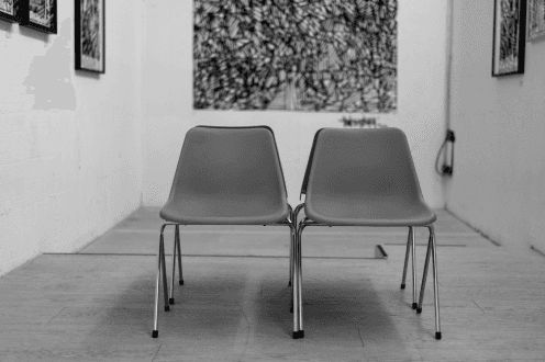 Two empty chairs symmetrically positioned in an art gallery, facing the viewer. This arrangement symbolizes the intersection of interdisciplinary research and artistic creation, inviting contemplation on innovative approaches to strategy development.