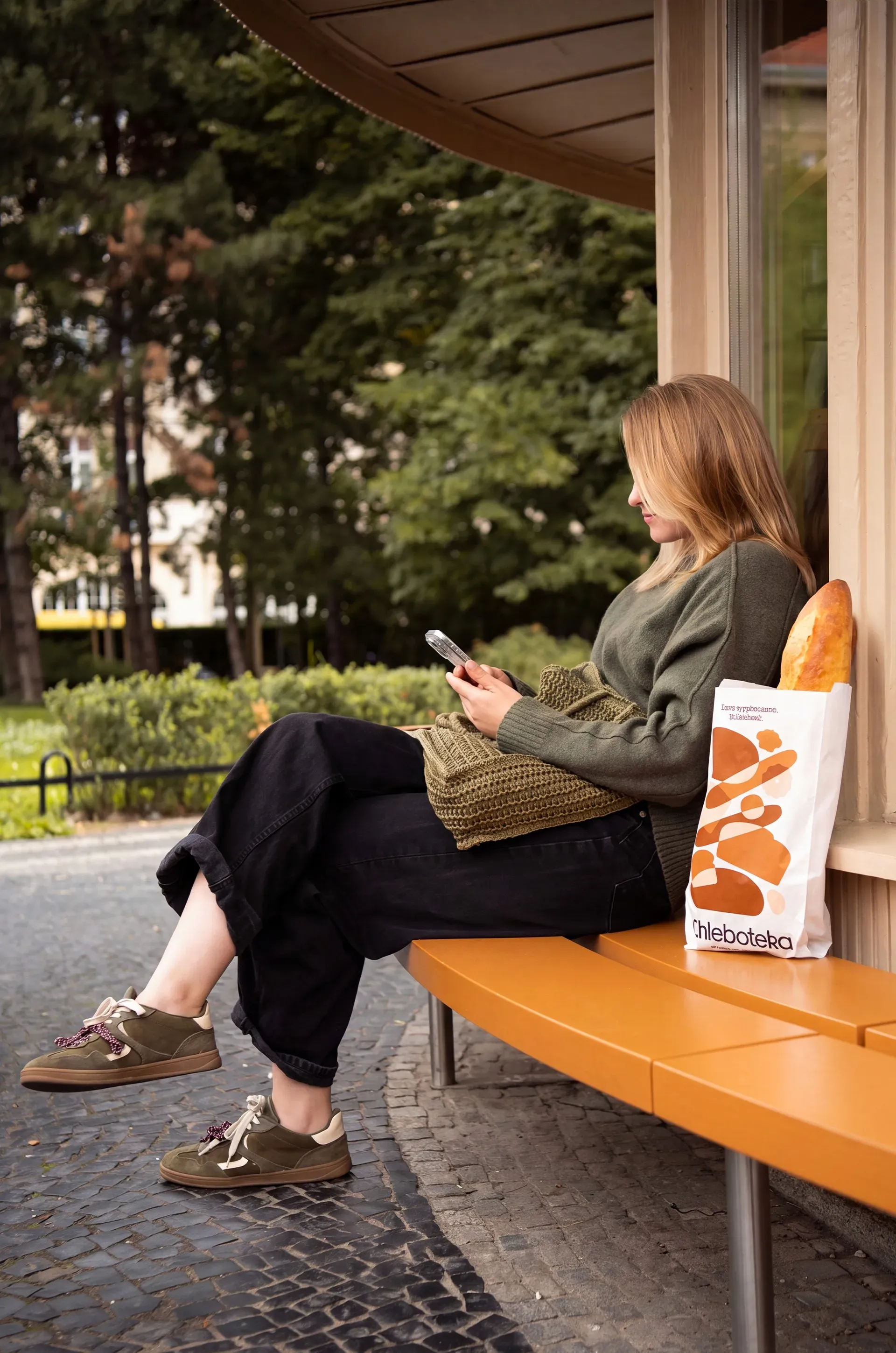 Woman on an orange bench with a Chleboteka bread bag and baguette, bakery packaging in use