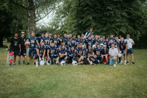 A large group of football players in blue and yellow uniforms pose together on a grassy field with trees in the background.