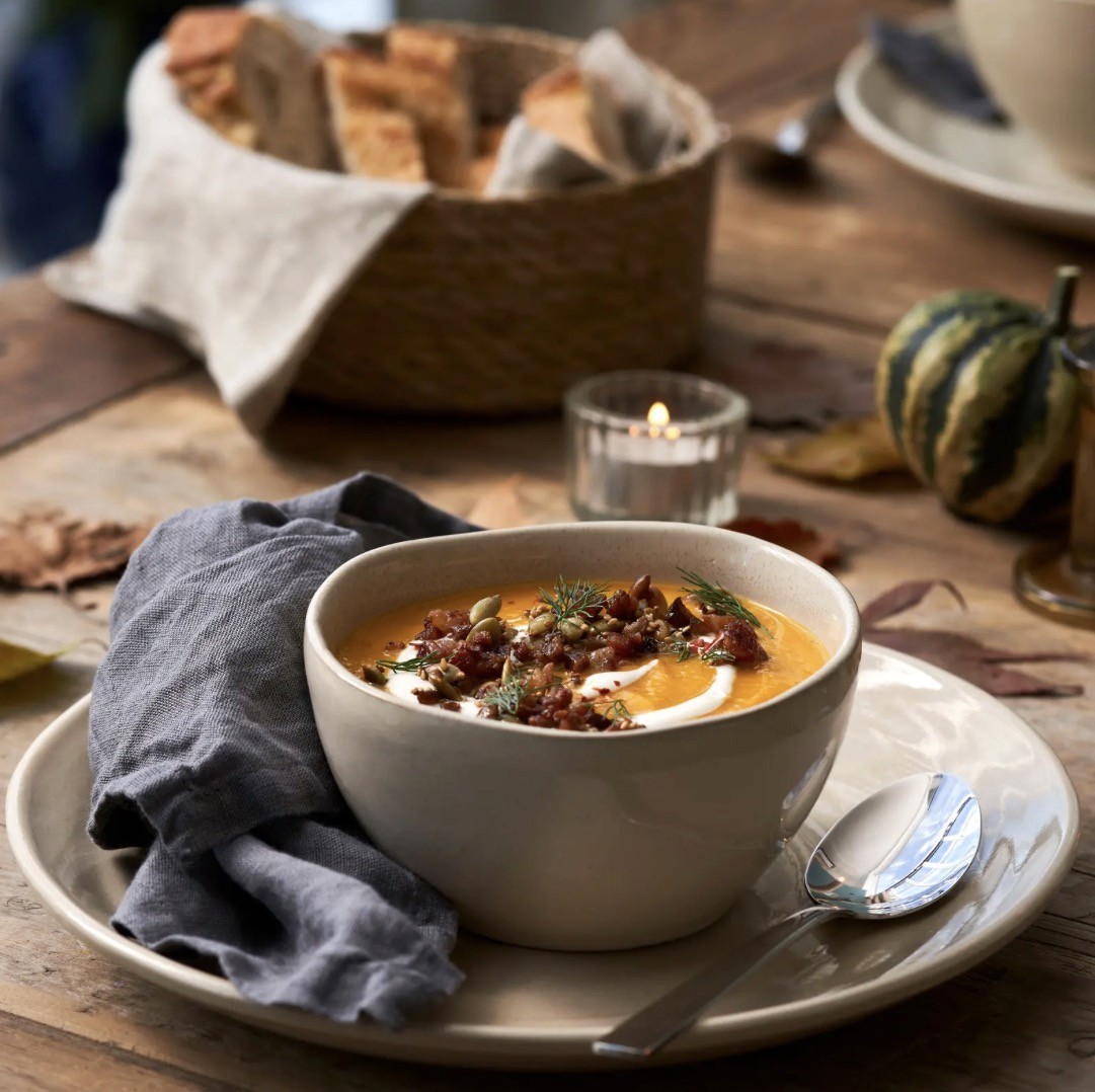 Bol en grès beige sur grande assiette, garni d'un velouté de potiron avec crème et garnitures, décor automnal avec courge et bougies