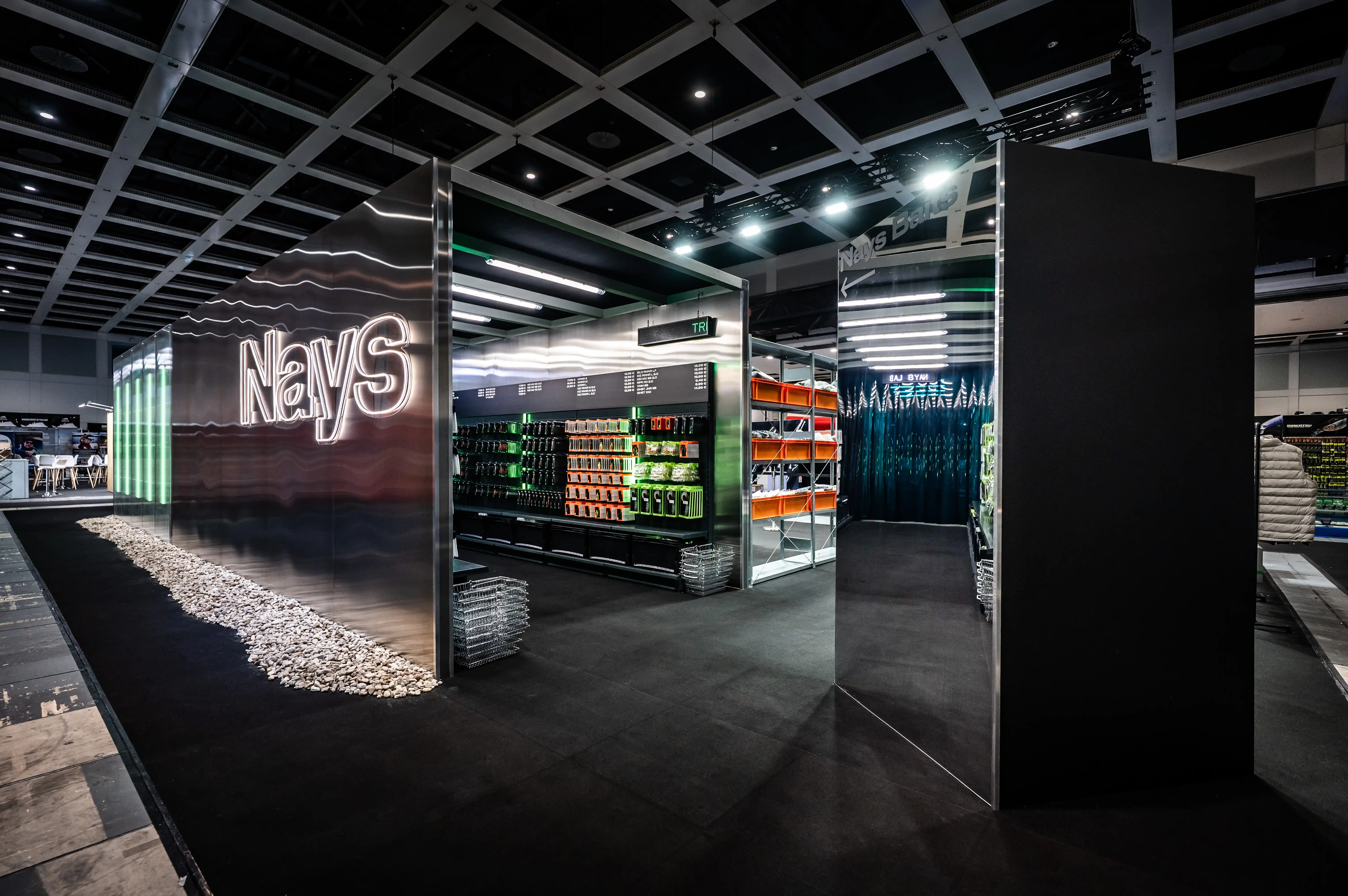 Entrance to the Nays booth with glowing logo on brushed metal wall, pebble bed and shelves of products.