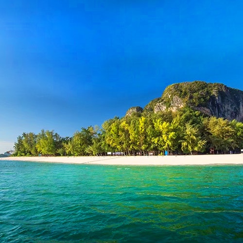 Clear turquoise waters meet a sandy beach lined with lush green trees and a rocky hill under a bright blue sky.