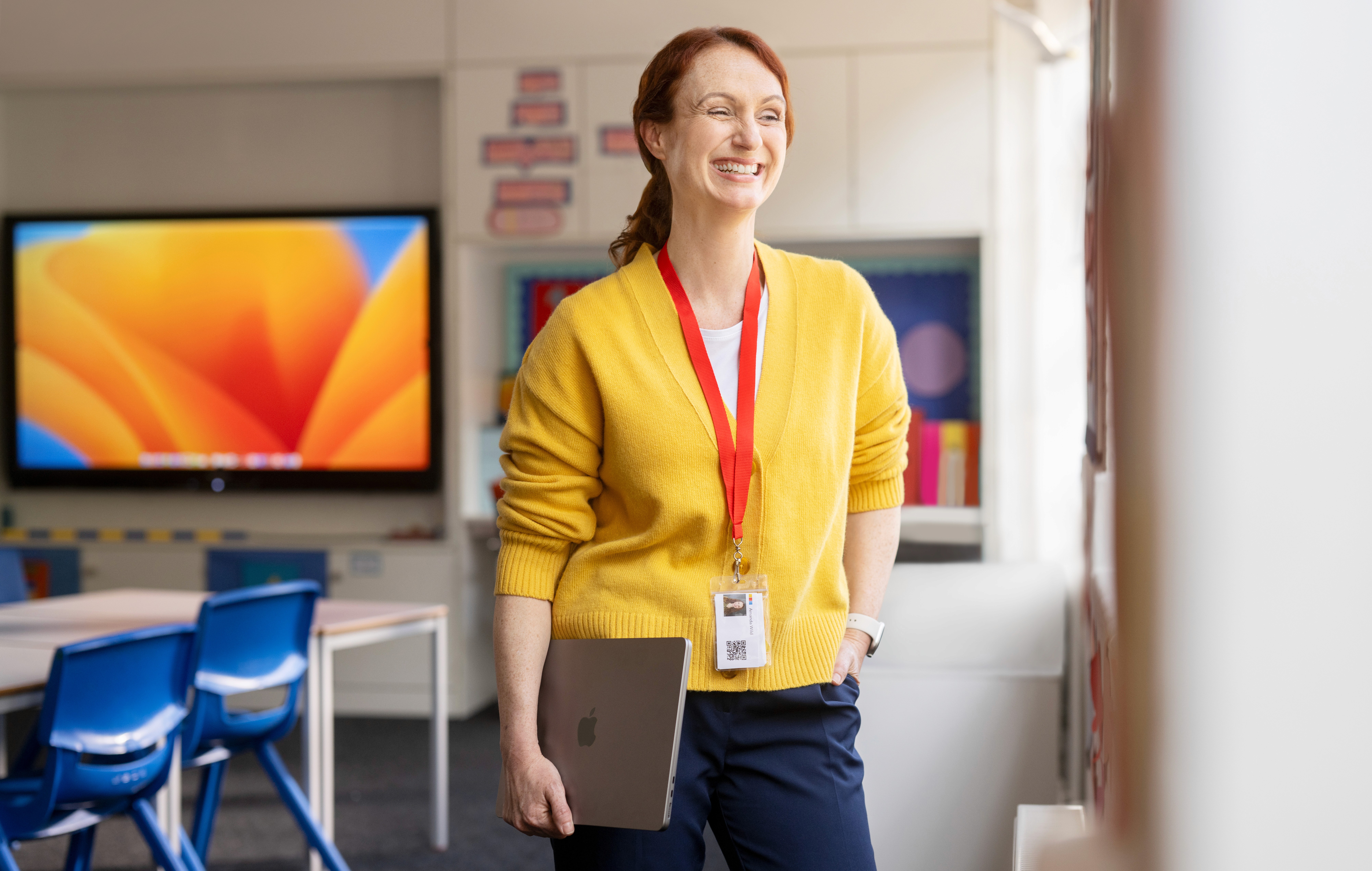 Lächelnde Lehrerin mit MacBook unter dem Arm in einem modernen Klassenzimmer mit iMac im Hintergrund