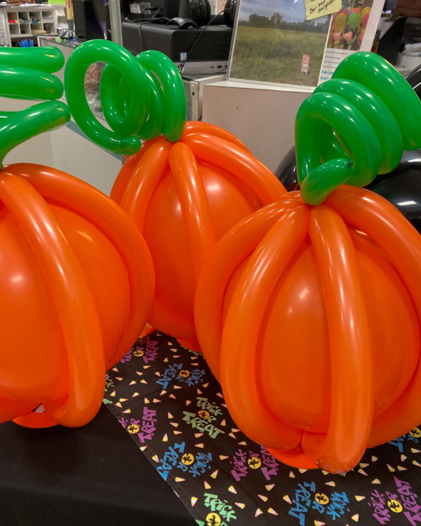 Three vibrant orange balloons are twisted into the shape of pumpkins, topped with green balloon stems, set against a festive Halloween-themed tablecloth.