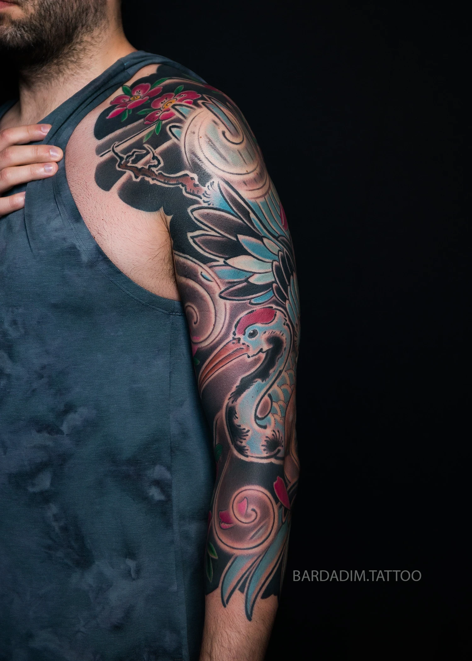 Outer view of the sleeve: the red-crowned crane's neck curves down the arm to the head near the elbow, layered blue wing feathers above and sakura at the shoulder.