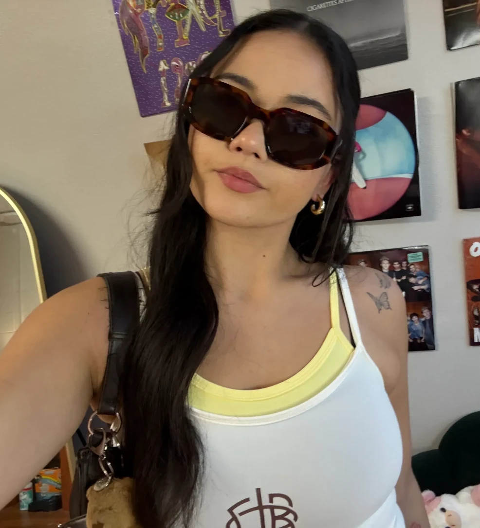 Indoor selfie of a person wearing oversized sunglasses and a white tank layered over a yellow top