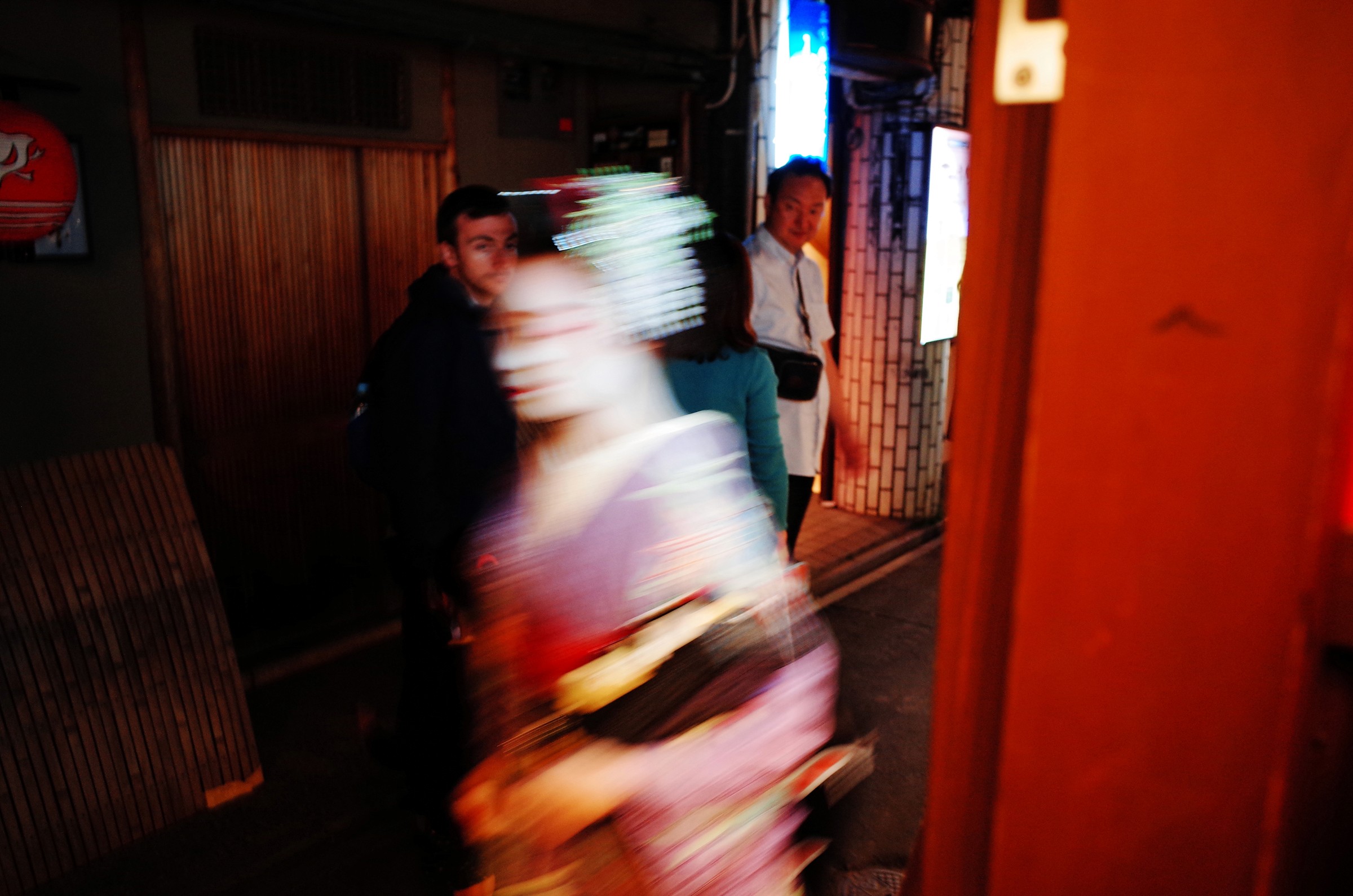 Photographie de rue à Kyoto d'une geisha en flou de mouvement passant furtivement dans une rue et deux touristes se retournant sur eux-mêmes pour la regarder