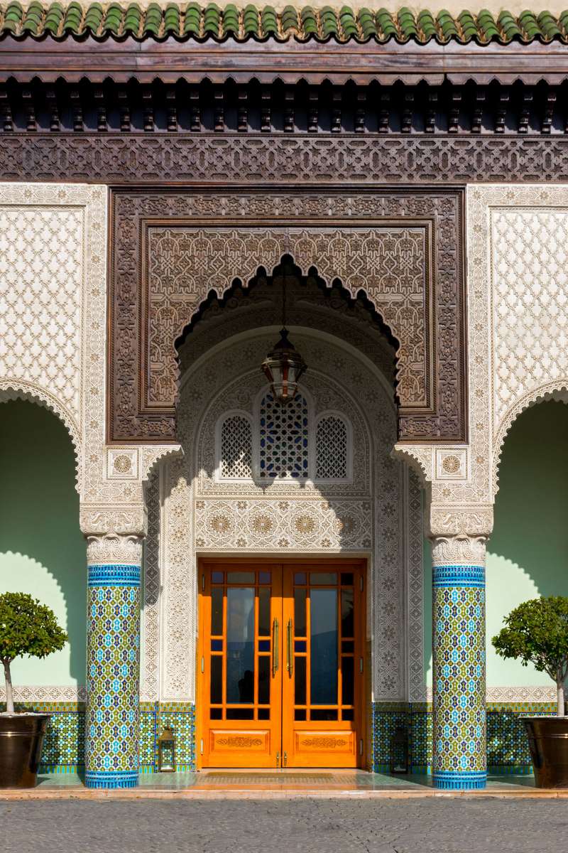 An ornate Moorish archway with intricate tilework and a carved wooden facade frames an orange door in a courtyard.