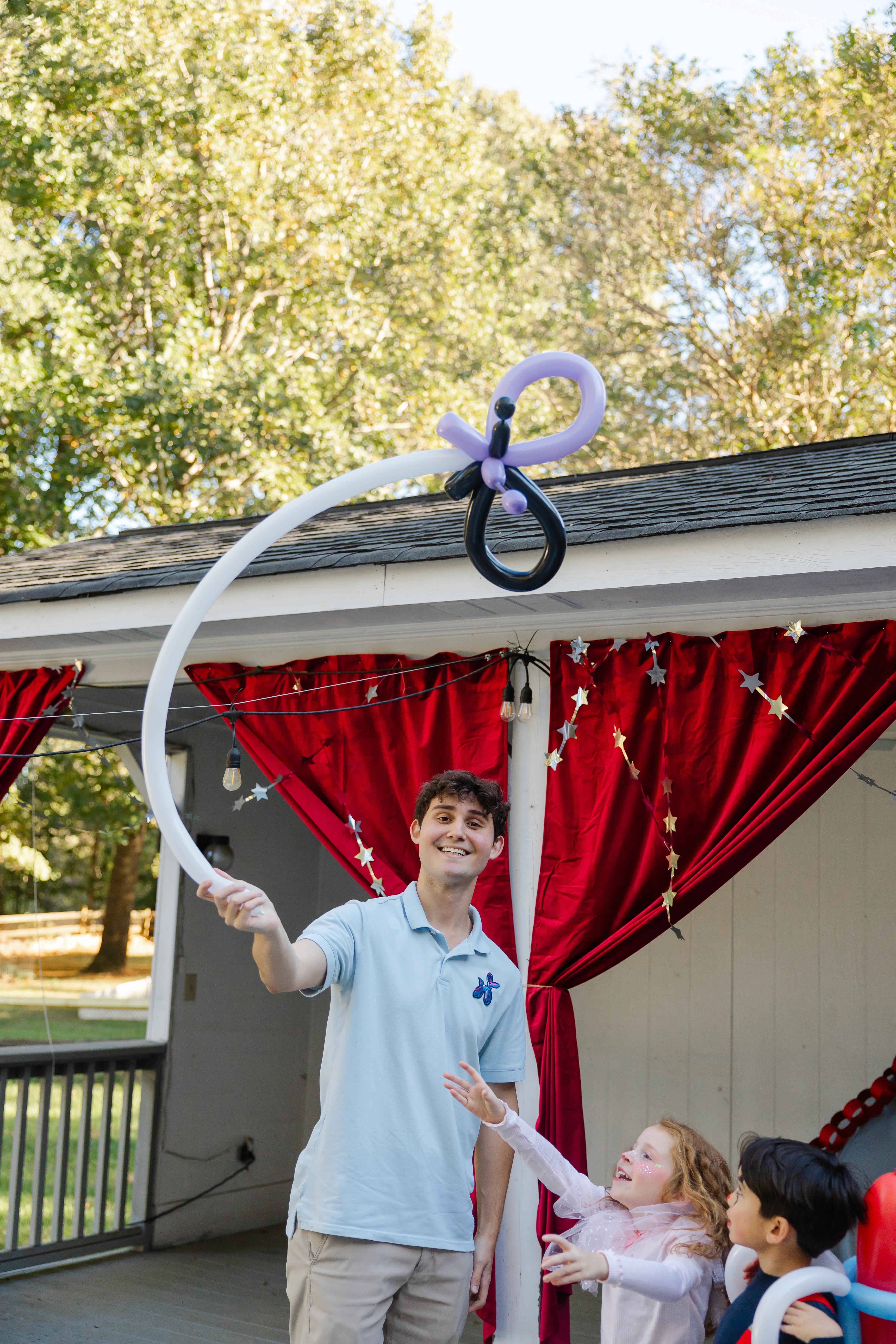 A balloon artist in a light blue shirt playfully holds up a large butterfly-shaped balloon as kids eagerly reach out for it. The setting is a backyard with red curtains, creating a lively and magical atmosphere.