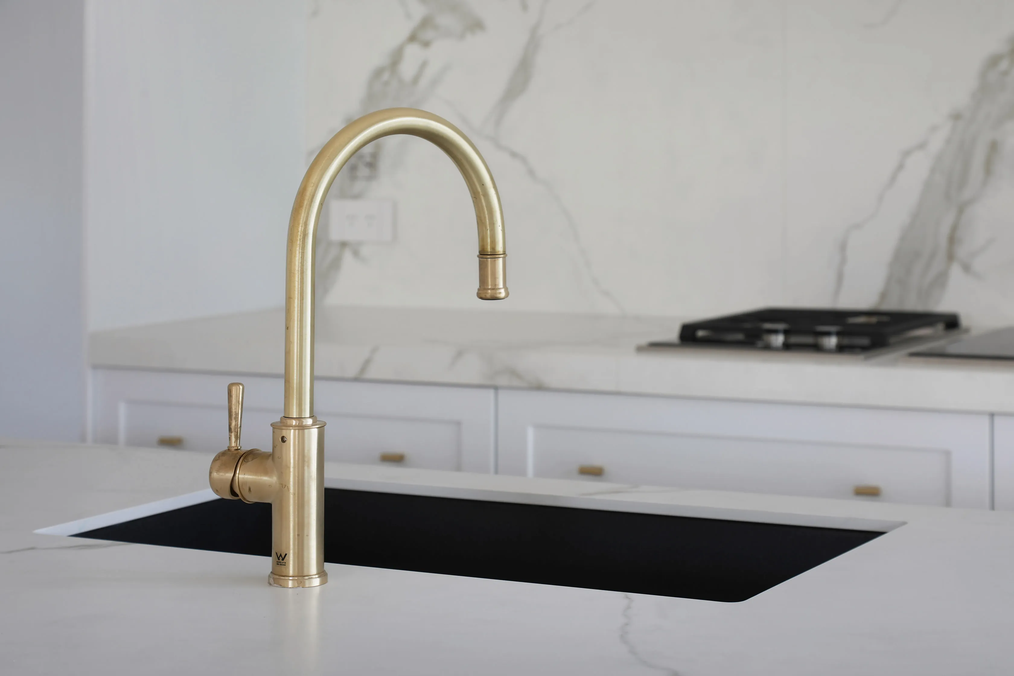 Kitchen island detail with a Calacatta-veined stone benchtop and splashback, an aged-brass gooseneck tap, black undermount sink and a gas cooktop in the background