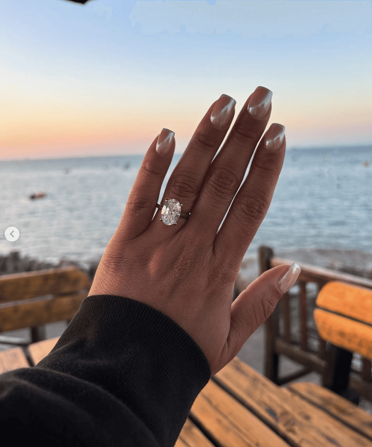 A couple stands on a sandy hill at sunset, facing away, with arms raised in a relaxed "hang loose" gesture. The scene conveys tranquility and joy.