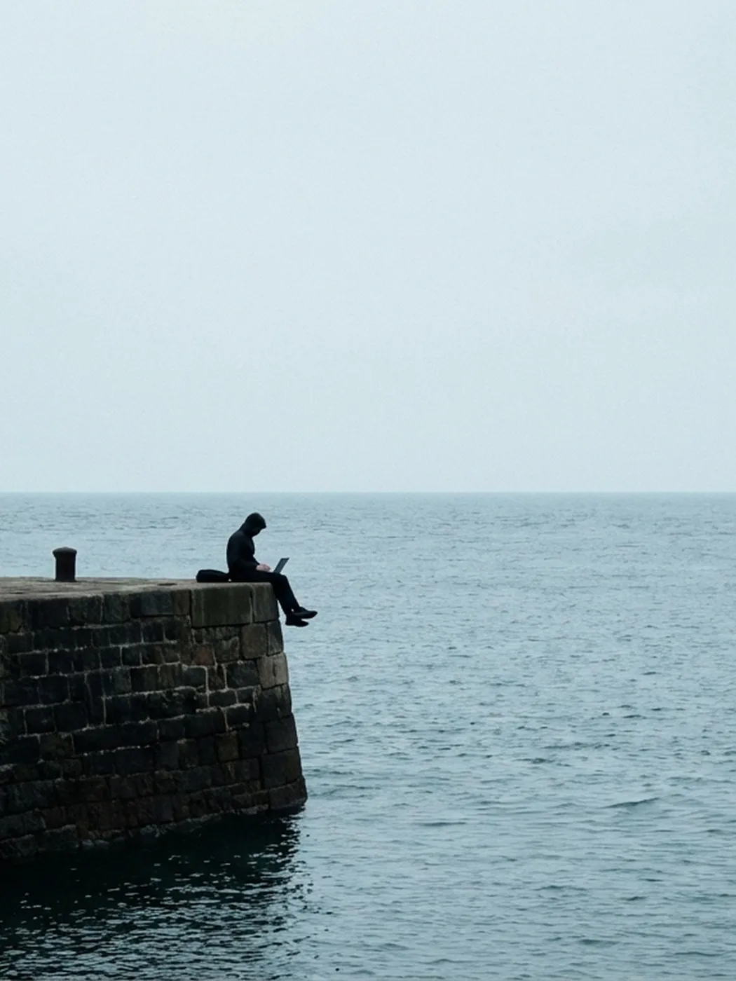 Figure with laptop sits on pier edge by the sea