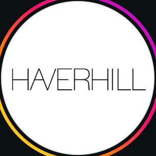 A woman with blonde bob hair in a grey coat and beige turtleneck stands against a neutral backdrop. She has a serious expression, conveying elegance.