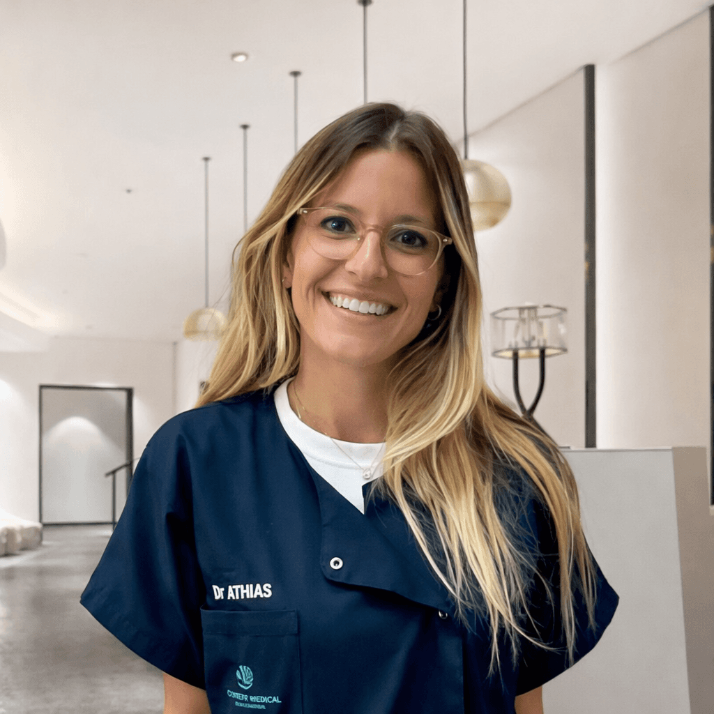 Close-up portrait of a young woman with green eyes, natural makeup, and dark hair, gently touching her face, against a neutral beige background