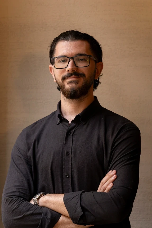 Smiling team member with glasses; crossed arms, soft indoor light, plain beige wall.