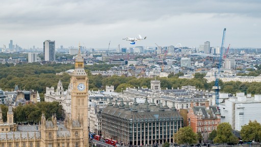 Aerial view of London featuring the iconic Big Ben, surrounded by historic architecture and lush green spaces, with modern city buildings and cranes in the background under a cloudy sky. A drone is flying through the sky, past Big Ben.