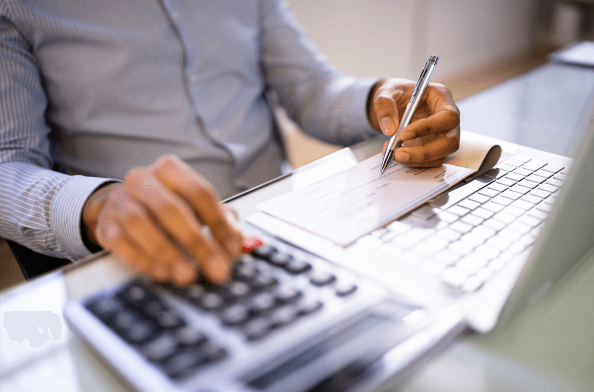 A person's hands using a calculator and writing on a document while working on a laptop.