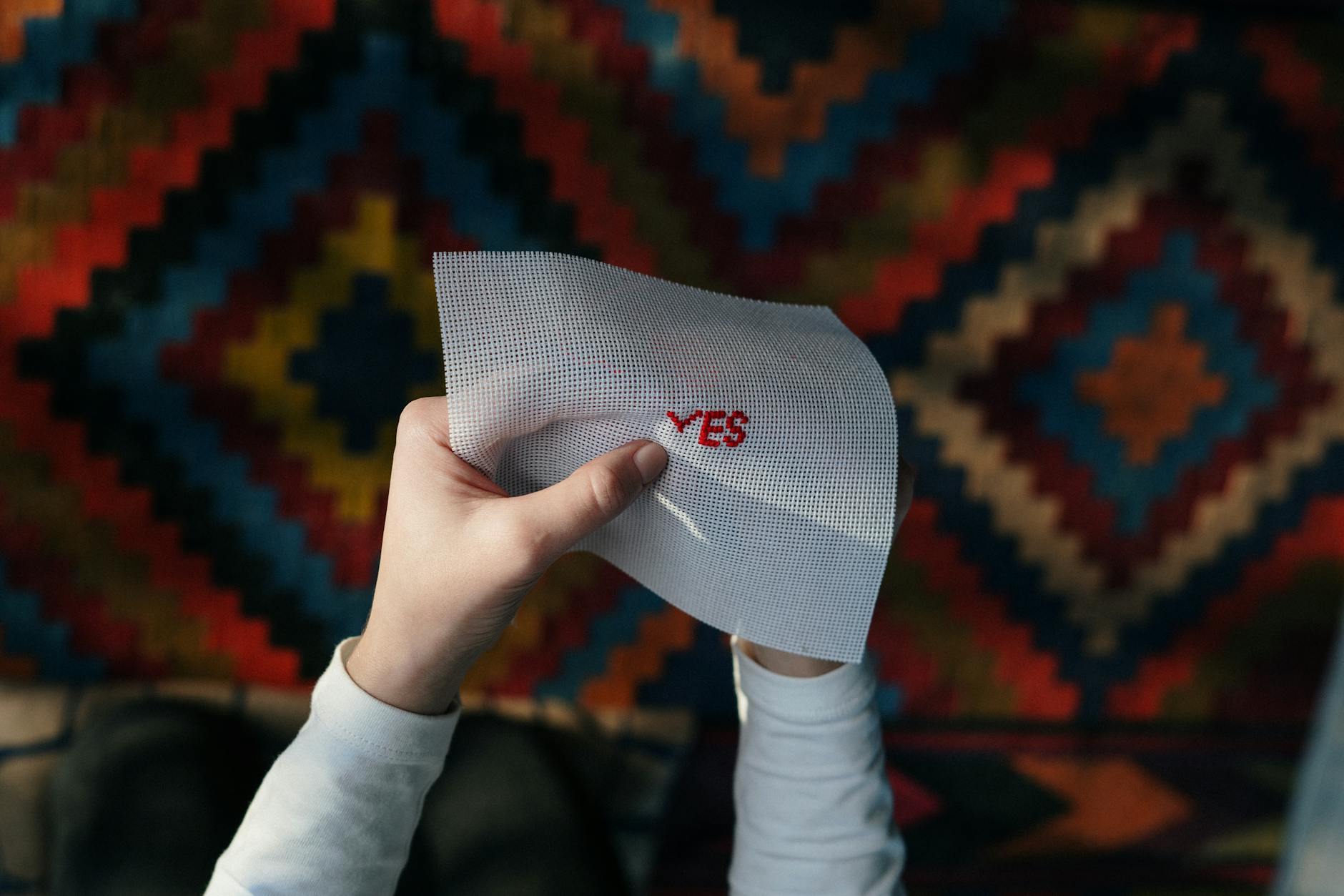 Hands holding an embroidered 'Yes' piece with colorful geometric background, showcasing needlework art.