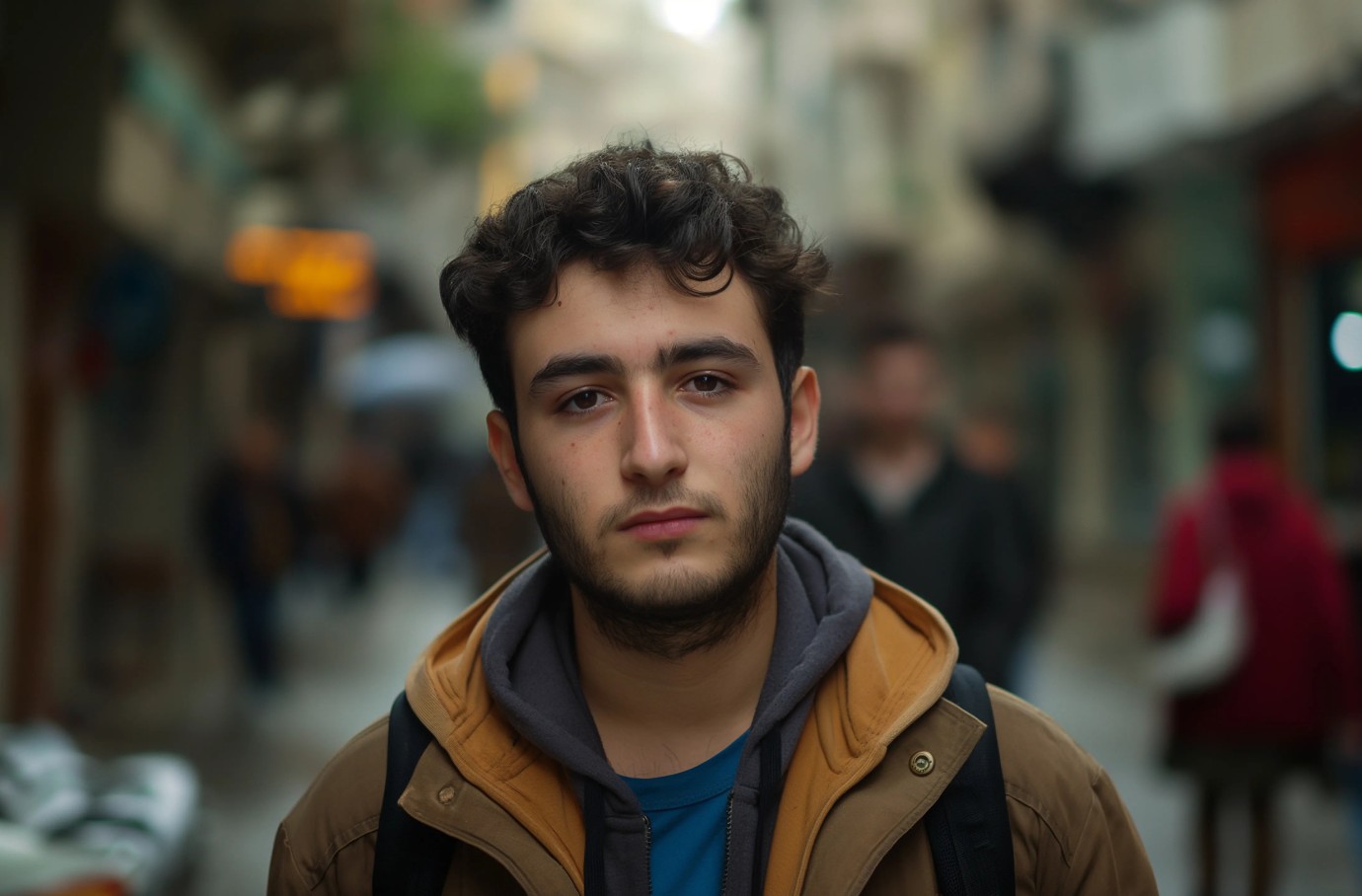 A young man with wavy dark hair and a beard stands on a bustling city street, wearing a brown jacket and backpack, with blurred pedestrians and buildings in the background.