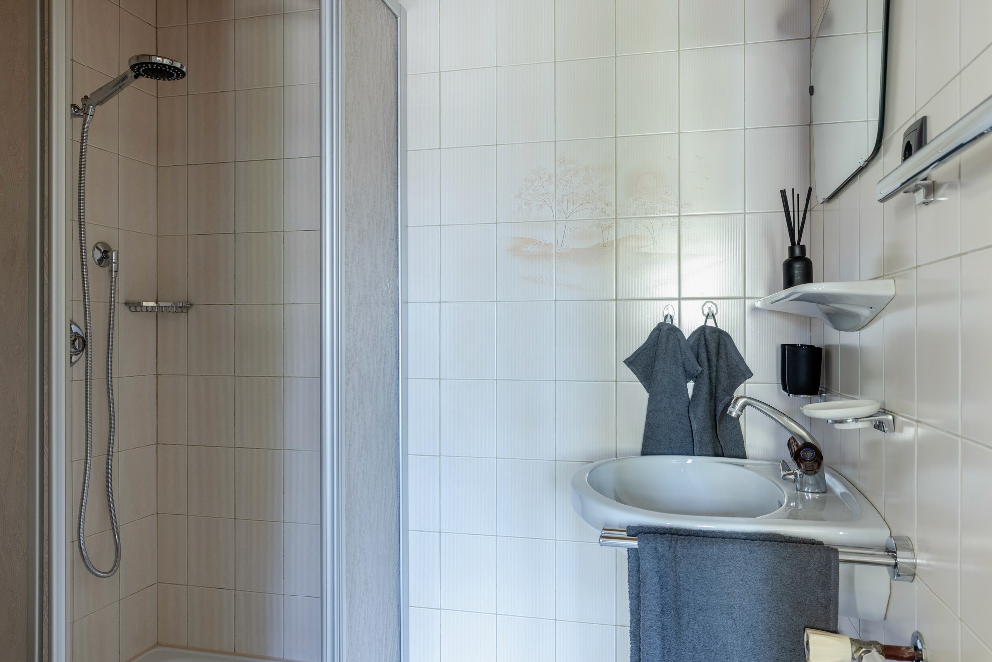 Ein modernes Badezimmer verfügt über eine ebenerdige Dusche mit einem eleganten Duschkopf aus Chrom, ein weißes Keramikwaschbecken mit einem dunklen Handtuch daran und minimalistische schwebende Regale mit Toilettenartikeln und Dekoration vor einer gefliesten Wand.
