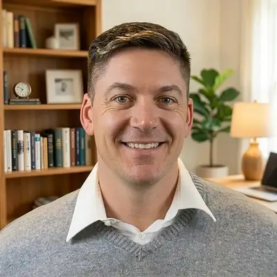 Profilbild eines lächelnden Mannes mittleren Alters mit kurzen dunkelbraunen Haaren, weißem Hemd und grauem Pullover. Im Hintergrund ein Bücherregal mit Büchern, einer Uhr und einer Pflanze sowie einer Stehlampe. Das Bild ist kreisförmig zugeschnitten mit hellblauem Rahmen.