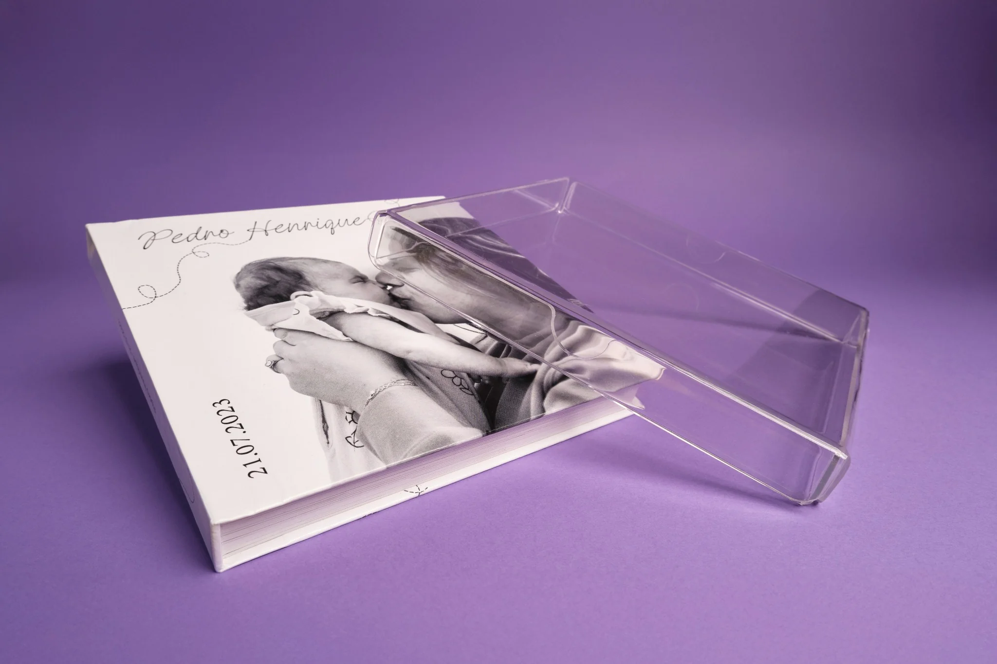 Mãos segurando fotolivro com capa de Arraial do Cabo sobre mesa de madeira clara, ao lado de flores cor-de-rosa e lilás