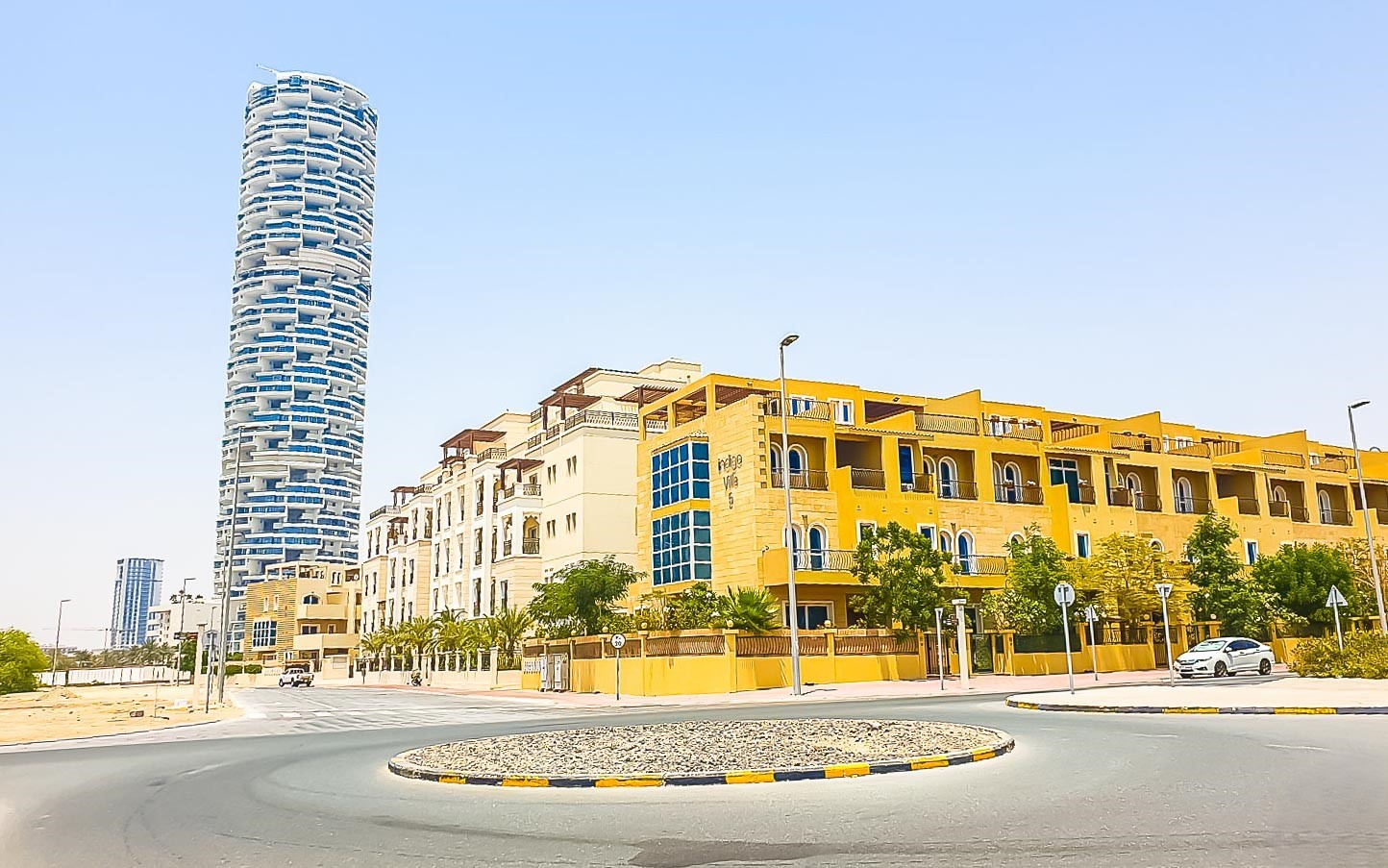 Street view in Jumeirah Village Circle featuring Mediterranean-style residential buildings in yellow and beige under a bright, clear sky.