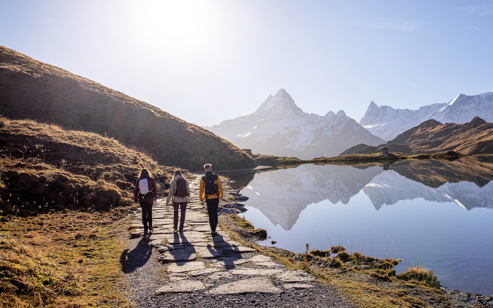 Hikers walking along a path by a lake with snow-capped mountains at Grindelwald First.