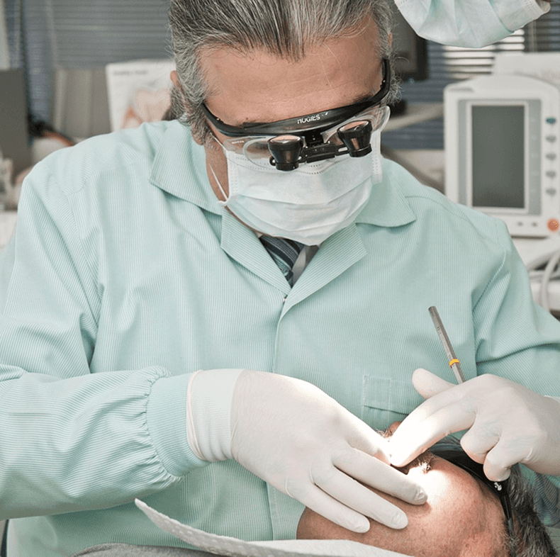 A dentist wearing magnifying glasses and a surgical mask performs a dental procedure on a patient in a clinic, surrounded by medical equipment.
