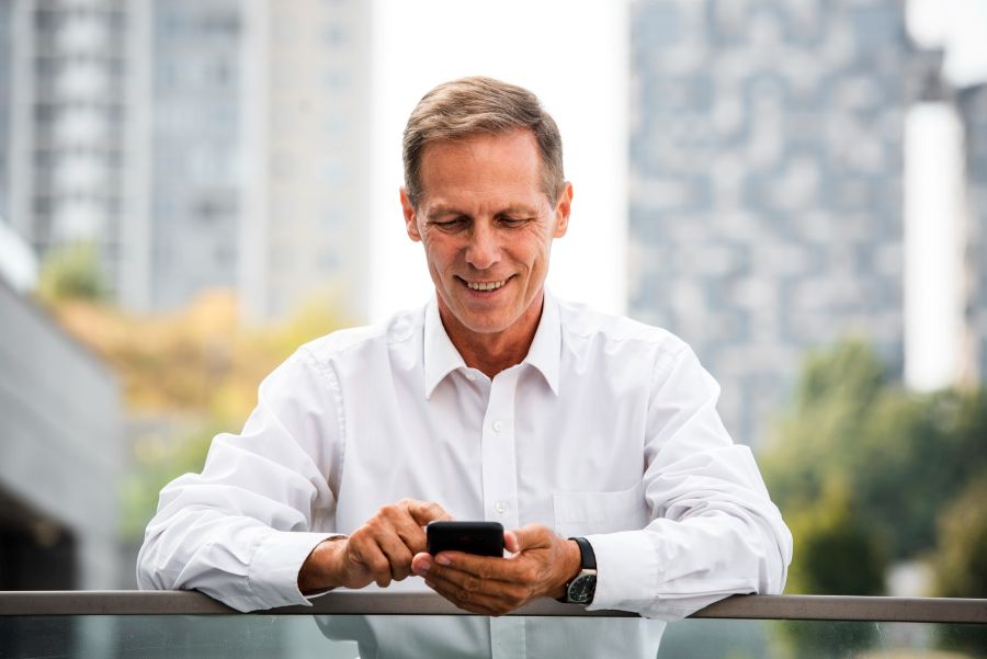 A middle-aged man in a white shirt is standing outdoors on a balcony, smiling and focused on his smartphone, with a blurred urban skyline in the background on a sunny day.